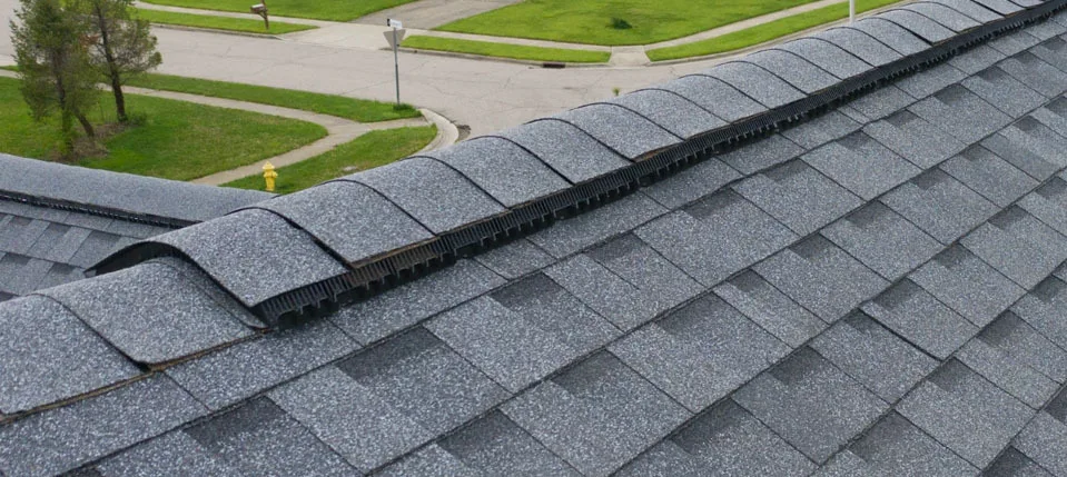 A continuous ridge vent running along a roof peak with the cap shingles partly lifted, showing the black vent material underneath that lets attic air escape